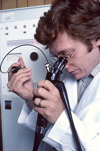A Caucasian male physician using a remotely controlled endoscope. He is looking through a microscope-like eyepiece to monitor his actions while using small brushes and knives to take a biopsy.
